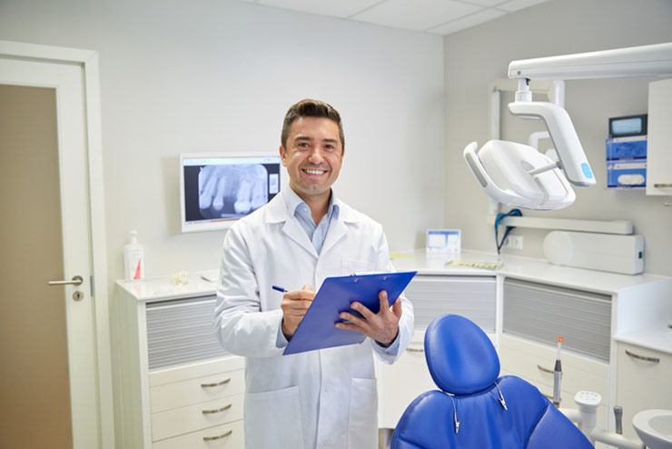 Smiling male doctor holding clip board and pen in exam room